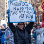A protester holds a large handmade sign that reads, "Protect Our Neighbors, Pass NY For All Act Now," written in bold black letters with a blue border. The demonstrator, wearing a black puffer jacket, red scarf, and a black beanie, raises the sign high while chanting. They wear gloves and glasses, appearing determined as they march down the street. Behind them, other participants hold various signs, including one that partially reads "LIBERTY." The setting is a city street lined with tall residential buildings, with the march progressing through a densely populated urban environment.
This protest is part of the 50 50 1 march in New York City on February 17, 2025, advocating for immigrant rights and passing the NY For All Act, which seeks to protect immigrant communities from local law enforcement cooperation with ICE. The atmosphere is passionate, with demonstrators calling for justice, safety, and equal rights for all New Yorkers.