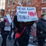 A demonstrator at the 50 50 1 march in New York City on February 17, 2025, holds up a bold, handwritten sign reading, "NEVER AGAIN MEANS NOW! PROTECT: WOMEN, DREAMERS, LGBTQ+, IMMIGRANTS, FAMILIES, FEDERAL WORKERS, ALL OF US!" The sign features a mix of black, red, blue, and green lettering, emphasizing urgency and inclusivity. The protester wears a black fuzzy jacket, black pants, and gloves, with a red crossbody bag adding a pop of color. Their face is obscured by the sign as they march in solidarity.
The background includes other protesters, some carrying newspapers with political messaging, while others hold additional signs demanding justice and protections for marginalized communities. The setting is a New York City street lined with red-brick buildings, with a crowd marching in support of human rights, immigrant protections, and equality.