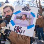 A demonstrator at the 50 50 1 march in New York City on February 17, 2025, holds a humorous protest sign featuring the phrase "NO COUP FOR YOU!" in bold black and red letters. The sign includes an image of Yev Kassem, the "Soup Nazi" from Seinfeld, taped onto the board with blue painter's tape. This clever reference humorously denounces attempts at political coups and authoritarianism.
The protester is wearing a brown work jacket, white gloves with black grips, and a backpack strap visible over their shoulder. They are holding multiple bottles, including a black "DORK" thermos and a green insulated bottle. They have long dark hair and a beard, giving a determined expression while marching with the crowd.
The background shows other participants in warm winter clothing, engaged in the demonstration. The sun shines brightly, casting natural light over the event, illuminating the peaceful but firm resistance against threats to democracy.