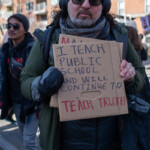 A demonstrator at the 50 50 1 march in New York City on February 17, 2025, stands holding a handmade cardboard sign reading, "I TEACH PUBLIC SCHOOL AND WILL CONTINUE TO TEACH TRUTH." The words "TEACH TRUTH" are written in bold red letters, emphasizing the commitment to education and factual instruction. The protester is bundled up for the cold weather, wearing a dark green jacket, a knit beanie, black sunglasses, and large over-ear headphones.
The background includes other marchers, some carrying protest signs, and residential buildings lining the street. The presence of a crowd suggests a strong turnout for the event, which appears to be focused on issues of education, democracy, and resistance to political censorship.
This image captures a moment of educator activism, highlighting the ongoing debate about academic freedom, curriculum control, and the role of teachers in shaping young minds. The determined expression on the protester’s face conveys a sense of defiance and dedication to truth in education.