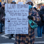 A demonstrator at the 50 50 1 march in New York City on February 17, 2025, holds a large handwritten sign titled "LAWS BROKEN," listing three U.S. laws:
The Privacy Act of 1974 – Protects personal information from federal agencies.
The Computer Fraud & Abuse Act – Criminalizes cyber-based attacks.
18 U.S. Code § 1752 – Prohibits trespassing on restricted government grounds.
The protester is bundled in a plaid coat with shades of blue, orange, and beige, a white knit beanie, sunglasses, and a blue face mask. They are also wearing black gloves and carry a crossbody bag with a colorful design featuring a sun and the word "SUN."
The background is filled with other demonstrators, some holding protest signs, and city buildings lining the street. The image captures a moment of civic engagement, emphasizing concerns about privacy rights, government accountability, and cyber-related regulations. The detailed sign suggests a protest focused on legal violations and justice.