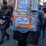 A demonstrator at the 50 50 1 march in New York City on February 17, 2025, holds up a handmade protest sign covered in duct tape and decorated with bold, handwritten messages. The sign reads:
"Don't scream about egg prices! Scream about our freedoms being stolen!"
"Don't roll over!"
"Join the Resistance! Before our country is UNRECOGNIZABLE!"
The sign incorporates real egg cartons taped to the board, reinforcing the metaphor about misplaced outrage. The protester is dressed in black, wearing black boots, and carrying a large black purse. Their face is mostly hidden behind the sign, focusing attention on the message.
In the background, other demonstrators march with protest signs, bundled in warm winter clothing. The atmosphere is charged with activism and urgency, as participants advocate for civic engagement and resistance against perceived political injustices.