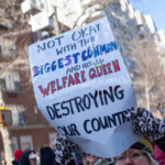 A protester at the 50 50 1 march in New York City on February 17, 2025, raises a boldly decorated sign with a strong political statement. The sign reads:
"NOT OKAY WITH THE BIGGEST CONMAN AND HIS WELFARE QUEEN"
"DESTROYING OUR COUNTRY"
The words "BIGGEST CONMAN" and "WELFARE QUEEN" are written in large, colorful letters, with the latter featuring a small crown illustration. The sign appears slightly crumpled, possibly from the wind or frequent handling.
The protester, an older woman, wears a black winter coat, a yellow scarf, and patterned gloves. Her facial expression conveys determination and concern. She stands firmly, holding the sign high with both hands.
In the background, fellow demonstrators march with additional protest signs, dressed in warm clothing. The urban setting, with tall apartment buildings and traffic lights, situates the demonstration in a busy city environment. The clear blue sky adds contrast to the scene, emphasizing the strong civic engagement of the participants.