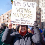 A protester smiles while holding a handmade sign reading "THIS IS WHY VOTING MATTERS, BUT YOU CAN STILL RAISE YOUR VOICE" at the 50 50 1 march on February 17, 2025, in New York City. The message emphasizes both the importance of electoral participation and continued activism beyond the ballot box.
The demonstrator is dressed in a navy winter coat, a red scarf, and black gloves, braving the cold weather while standing among a diverse crowd of protesters. In the background, other demonstrators can be seen carrying signs, and a traffic light glows green, symbolizing movement and progress. This moment captures the energy of civic engagement and the power of collective action.