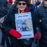 An older protester, dressed in a black winter coat, black beret, red scarf, and red gloves, holds a satirical sign reading "EVIL CHILD, DEMON DAD." The sign features a caricature of Donald Trump and Elon Musk, emphasizing political criticism. She wears sunglasses and a yellow lanyard, standing firmly with a black crossbody bag and a walking cane, participating in the 50 50 1 march in New York City on February 17, 2025.
The background captures a crowd of demonstrators in heavy winter clothing, many wearing hats and scarves to keep warm. Their presence signifies the broader political activism and civic engagement of the event, where protesters voiced concerns about democracy, corporate influence, and social justice.
This image highlights the intersection of humor and resistance, as demonstrators use creative visuals to amplify their messages in a peaceful protest setting.