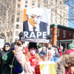 During the 50 50 1 march on February 17, 2025, in New York City, a protester raises a striking sign featuring an illustrated image of Donald Trump with the word "RAPE" in bold white letters. Below the word, a website URL, "weare.sigh.today," is visible. The protester, dressed in a pink faux fur coat, black knit hat, red scarf, and pink face mask, holds the sign high while gripping a dog leash with a small dog by her side. The crowd behind her, clad in winter clothing, holds various signs advocating for justice, women's rights, and political accountability.
The demonstration, held in Washington Square Park, was part of a larger pro-democracy and social justice movement, with participants condemning political corruption, authoritarianism, and violations of human rights.