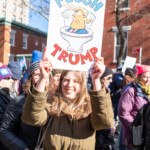 A young protester smiles while holding a bold sign that reads "FLUSH TRUMP," featuring a cartoon image of Donald Trump’s head inside a toilet. She wears a green parka and carries a canvas tote bag over her shoulder. The 50 50 1 march in New York City on February 17, 2025, drew a large crowd advocating for democracy, accountability, and political change. Surrounding her, other protesters, dressed in winter clothing, hold signs with various political messages. The event took place in Washington Square Park, a historic site for activism and demonstrations.