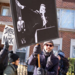 A protester in a navy coat, black gloves, and a knit beanie holds a striking black-and-white sign depicting Elon Musk as a puppet master controlling Donald Trump. The image on the sign is styled like propaganda art, with Musk appearing in a dictatorial pose while manipulating Trump with strings. The 50 50 1 march, held on February 17, 2025, in New York City’s Washington Square Park, brought together demonstrators advocating for democracy, accountability, and resistance against authoritarianism. Other participants in the background hold protest signs, including one that reads “IMPEACH PRES. MUSK.” The crowd, dressed for winter, engages in discussions and activism, reinforcing the protest’s strong political messages.