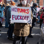 A protester at the 50 50 1 march in New York City holds a bold sign reading “NAZI PUNKS F* OFF”**, a reference to the Dead Kennedys' 1981 anti-fascist punk anthem. The individual wears a white face mask, sunglasses, an olive green parka, black cropped pants, and high-top sneakers, standing amidst a crowd of demonstrators. The protest, which took place on February 17, 2025, in Washington Square Park, saw activists rallying against authoritarianism, white nationalism, and far-right extremism. Other protesters in the background carry signs and engage in conversations, reinforcing the event’s atmosphere of resistance and solidarity.
