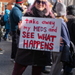 A protester with pink hair, cat-eye sunglasses, and a black coat stands confidently at the 50 50 1 march in New York City, holding a sign that reads “Take away my MEDS and SEE WHAT HAPPENS” in bold red and black letters with a small smiley face. The message is a powerful statement on the importance of access to mental health medication. The event, which took place on February 17, 2025, in Washington Square Park, gathered activists advocating for human rights, healthcare access, and social justice. Behind her, a crowd of demonstrators holds various signs, emphasizing the collective call for justice and policy change. The protester’s stance and expression convey both defiance and determination, reinforcing the urgency of the message.