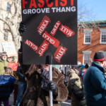A protester in a brown fur coat, black gloves, and a scarf holds up a bold black sign with white and red text at the 50 50 1 march in New York City on February 17, 2025. The sign reads: “WE DON'T NEED THIS FASCIST GROOVE THANG”, referencing the 1981 song by Heaven 17, with a design incorporating torn-up "Make America Great Again" slogans. The message condemns far-right ideology and critiques authoritarian tendencies in politics. The protest took place in Washington Square Park, drawing activists and community members advocating for democracy, human rights, and social justice. The background shows a diverse crowd carrying various protest signs, reinforcing the collective resistance against extremism.