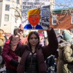 A young woman with long dark hair, a brown cropped puffer jacket, a black top, blue jeans, and a gold chain belt holds up a powerful sign at the 50 50 1 march in Washington Square Park, New York City, on February 17, 2025. Her sign, featuring an exaggerated caricature of Donald Trump with an orange face and yellow hair, reads: “YOUR GROCERY PRICE ISN’T MORE IMPORTANT THAN MY HARD-WORKING IMMIGRANT MOM!” Additional handwritten messages on the sign include “Fuk Trump,” “Let Us Be Kids,” and “Fuck the Oligarchy.” This statement highlights the struggles of immigrant families in the face of economic and political challenges. She stands confidently in the midst of a diverse crowd of protesters advocating for immigrant rights, democracy, and social justice. Other demonstrators holding signs are visible in the background, reinforcing the atmosphere of activism and community solidarity.