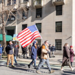 A group of protesters walks down the street during the 50 50 1 march in New York City on February 17, 2025. One man in the center, wearing a brown jacket, sunglasses, and gloves, proudly waves a large American flag as he marches with fellow demonstrators. Another protester behind him holds a partially visible sign that reads “LESS”, hinting at a message about democracy or social justice. The group, dressed in warm winter clothing, strides past a city building with a green awning that reads “One Fifth Avenue”, a well-known address in Manhattan. Police officers stand in the background observing the march. The atmosphere reflects peaceful activism, patriotism, and civic engagement as the demonstrators advocate for their cause.