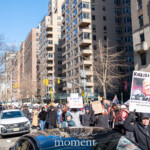 A large crowd of demonstrators takes to the streets of New York City on February 17, 2025, as part of the 50 50 1 march, advocating for democracy and against authoritarianism. In the foreground, a protester holds a striking sign reading “Kakistocracy”, defining it as "government by the least qualified or most unprincipled citizens." The sign features an image of Donald Trump, emphasizing the protest’s critical stance on governance. Other participants carry signs with messages such as “We Will Not Uphold Tyranny” and “Stand & Fight Back”, reinforcing the theme of civic resistance. The demonstrators, dressed for winter, move down a wide Manhattan avenue lined with tall residential buildings, with traffic lights and city signs visible in the background. Their march symbolizes a collective call for action in a politically charged moment.