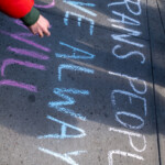 A powerful message is written in colorful chalk on a city sidewalk during the 50 50 1 march in New York City on February 17, 2025. The message, in white, blue, and pink chalk, reads “Trans People Have Always”, partially cut off but still resonating with a strong call for trans visibility and rights. A passerby in a red jacket reaches toward the artwork, emphasizing the interactive and communal nature of the demonstration. The use of trans flag colors in the lettering highlights the march’s support for LGBTQ+ rights amid a larger movement advocating for democracy, equality, and resistance against oppression.