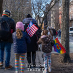 A family walks together during the 50 50 1 march in New York City on February 17, 2025, carrying symbols of both patriotism and inclusivity. Two young girls, dressed warmly in puffer jackets and colorful leggings, carry small flags—one holding an American flag, while the other waves a rainbow flag featuring a heart. An adult, dressed in a black coat with a rainbow scarf, walks alongside them, symbolizing a commitment to democracy and LGBTQ+ rights. The march moves through a city street lined with brownstone buildings, parked cars, and fellow demonstrators, emphasizing the intergenerational nature of the protest. This image captures a powerful moment of civic engagement, highlighting the role of families in activism.