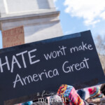 A protester holds up a black sign with bold white lettering stating, "HATE won’t make America Great" during the 50 50 1 march in New York City on February 17, 2025. The sign is secured with black tape at the corners, reinforcing its message against division and discrimination. A colorful knit glove peeks into the frame, adding a touch of vibrancy to the image. In the background, another sign partially visible reads, "Deport these 1% Musk, Melania," while the iconic Washington Square Arch stands tall in the distance, symbolizing resistance and activism in the heart of the city. The crowd is made up of demonstrators wearing warm clothing, indicating the event took place on a cold winter day. This moment captures the energy of the movement, emphasizing messages of unity and rejection of hate in America’s political discourse.