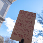 A protester holds up a handmade cardboard sign reading, "DEPORT THESE 1%: MUSK, MELANIA, VANCE’S WIFE, KASH PATEL, MEHMET OZ" during the 50 50 1 march in New York City on February 17, 2025. The message, written in bold red letters, is directed at a group of prominent individuals, reflecting strong political sentiments among the demonstrators. A gloved hand grips the sign tightly, raising it high against the backdrop of a bright blue sky and the towering Washington Square Arch, a historic landmark often associated with activism and resistance. Other protest signs are partially visible below, including one with the words "TRUE PATRIOT." The image captures the intensity and engagement of the marchers as they voice their opinions in a public demonstration.