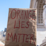 A protester holds up a handmade sign made from brown cardboard with the bold red message "ORANGE LIES MATTER" during the 50 50 1 march in New York City on February 17, 2025. The phrase is a play on words, seemingly criticizing former President Donald Trump, often nicknamed "Orange" due to his distinctive tan. The sign is lifted high against the backdrop of the Washington Square Arch, a well-known site for political activism. The crowd of demonstrators can be seen in the background, holding various signs as they march in unity. A protester wearing a gray "New York" beanie is partially visible in the foreground, adding to the dynamic energy of the demonstration. The bright blue sky and natural light illuminate the scene, highlighting the sign and its message.