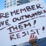 A protester holds a large white sign with bold, hand-painted lettering in black and red that reads: "REMEMBER WE OUTNUMBER THEM! RESIST!" The sign is raised high above the demonstrator’s head, with a gloved hand gripping the bottom edge. The background features a clear blue sky and city buildings, indicating an urban setting, likely New York City. This image captures the 50 50 1 march on February 17, 2025, a political demonstration advocating for democracy, resistance against authoritarianism, and collective action. The sign’s powerful message emphasizes the strength of numbers and urges continued civic engagement and defiance.