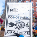 A protester holds up a striking black-and-white sign featuring a graphic of small fish uniting to overpower a larger fish, accompanied by the bold text: "DO NOT PANIC, ORGANIZE." The sign conveys a message of collective action and grassroots mobilization, encouraging people to stay calm and work together for change. The image was taken at the 50 50 1 march in New York City on February 17, 2025, a demonstration advocating for democracy, social justice, and resistance against authoritarianism. The sign is laminated for durability, and the protester wearing a blue jacket and gloves grips its corner firmly. The urban backdrop includes buildings and fellow demonstrators, reinforcing the scale and energy of the march.