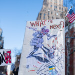 A protester at the 50 50 1 march in New York City on February 17, 2025, holds up a laminated sign featuring a political cartoon and the bold red text "WHAT'S NEXT?" The artwork depicts a figure dressed in Nazi-inspired military attire stomping on democratic institutions and services, with red ink resembling blood splatters around the edges. Various government programs, including USDA, Medicare, Medicaid, USAID, Veterans Affairs, and the Department of Energy, are listed, seemingly crushed under the authoritarian march. The sign suggests a warning about the erosion of democracy and public services under extremist policies. An American flag is attached to the sign’s corner, fluttering in the wind. The background includes a tall Art Deco-style building, other demonstrators, and additional protest signs, adding to the atmosphere of civic resistance.