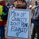 A protester at the 50 50 1 march in New York City on February 17, 2025, stands holding a boldly framed sign with a rainbow glitter border that reads: "MEN OF QUALITY DON'T FEAR EQUALITY" with a gender equality symbol drawn underneath. The person, wearing a black baseball cap with a rainbow heart emblem, sunglasses, and a thick scarf, looks directly at the camera with a calm yet determined expression. The background is filled with other demonstrators, some wearing high-visibility vests and warm winter clothing, indicating the event's organized and inclusive atmosphere. The setting is outdoors in Washington Square Park, where people have gathered to advocate for social justice, gender equality, and human rights.