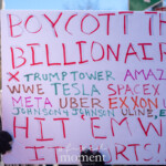 A protester at the 50 50 1 march in New York City on February 17, 2025, holds a large handmade sign reading "BOYCOTT THE BILLIONAIRES" in bold red letters. The sign lists major corporations and entities such as Trump Tower, Amazon, WWE, Tesla, SpaceX, Boeing, Meta, Uber, Exxon, Johnson & Johnson, and Uline, with the phrase "HIT 'EM WHERE IT HURTS!!" at the bottom, encouraging economic activism. The demonstrator, wearing black gloves and a winter coat, grips the sign firmly, emphasizing the protest's stance against corporate power and wealth concentration. Other demonstrators and protest signs are visible in the background, reinforcing the collective action of the movement. The event takes place in Washington Square Park, a historic site for political activism and social movements.