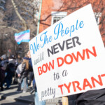 A protester at the 50 50 1 march in New York City on February 17, 2025, holds a large sign that reads: "We The People will NEVER BOW DOWN to a petty TYRANT" in a mix of bold black, blue, and red lettering. The phrase emphasizes defiance and a commitment to democracy. In the background, several transgender pride flags and rainbow heart flags are visible, held by other demonstrators marching through the streets near Washington Square Park. Protesters wear winter clothing, and some have helmets or masks, indicating a prepared and organized demonstration. The scene captures the energy of political activism and resistance against authoritarianism, with a diverse crowd advocating for democracy and social justice.