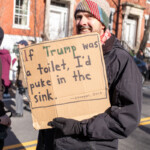 A demonstrator at the 50 50 1 march in New York City on February 17, 2025, holds up a handmade cardboard sign with a provocative and humorous message: "If Trump was a toilet, I'd puke in the sink. —Chopper, 2013". The protester, a bearded man wearing a colorful knit beanie, black gloves, and a winter jacket, smiles as he engages with the event. A camera pouch is attached to his belt, suggesting he may also be documenting the protest. The background features historic brick buildings and other protesters, some carrying cameras, adding to the lively and expressive atmosphere of the demonstration. This moment captures the mix of political critique and humor often found in activist movements.
