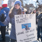 A young woman participating in the 50 50 1 march in New York City on February 17, 2025, holds a sign addressing Elon Musk’s alleged ableism. The sign reads: "HATE IS NOT AN AUTISTIC TRAIT! SHAME ON MUSK! Sincerely, The Neurodivergent Community", accompanied by an edited image of Musk at a presidential podium. She is dressed in a light blue puffer jacket and a blue beanie with a visible patch that reads "PATAGONIA TRADER JOE'S WASHINGTON D.C." She raises her left hand in a gesture of acknowledgment while standing among a crowd of demonstrators. Other signs and participants can be seen in the background, along with Washington Square Park’s black lampposts and trees, indicating the iconic location of the protest.