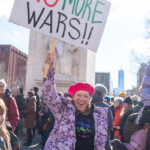 A woman enthusiastically raises a sign reading "NO MORE WARS!!" in bold red, black, and green letters at the 50 50 1 march in New York City on February 17, 2025. She is wearing a bright pink beret, clear glasses, and a black hoodie with neon-colored text that reads "FIERCE FULL Authentic" under a purple floral puffer jacket. Her broad smile radiates energy as she stands in front of Washington Square Park’s arch, with a crowd of protesters visible in the background, along with One World Trade Center in the distance. A trans flag is seen in the bottom right corner, adding to the diverse representation at the event.