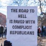 A protester at the 50 50 1 march in New York City on February 17, 2025, holds a large white sign with bold black text that reads: "THE ROAD TO HELL IS PAVED WITH COMPLICIT REPUBLICANS." Their face is obscured behind the sign, but they are dressed in a black puffer jacket and black gloves, with a colorful patterned bag strap visible on their left side. The background shows other demonstrators and leafless trees, with sunlight filtering through the branches, highlighting the wintry atmosphere of the rally.
