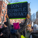 A protester at the 50 50 1 march in New York City on February 17, 2025, raises a large black sign with white text reading: "DON'T DISMANTLE DEMOCRACY." The sign is framed with a neon green border. The protester is wearing a dark puffer jacket, a bright pink knitted hat, brown gloves, and a crossbody bag. The background includes other demonstrators, some engaged in conversation, holding their own protest signs. A woman with curly hair, glasses, and a brown beanie is visible to the right, appearing to be in mid-discussion. The scene is set against a backdrop of red brick buildings and leafless trees, emphasizing the winter setting of the protest.