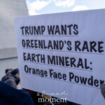 A close-up of a protest sign at the 50 50 1 march in New York City on February 17, 2025, features bold black text on a white background that reads: "TRUMP WANTS GREENLAND’S RARE EARTH MINERAL: Orange Face Powder." The sign satirically references former President Donald Trump’s past interest in purchasing Greenland, with a humorous jab at his signature orange-toned complexion. A protester in a dark winter coat and gloves holds the sign, while other demonstrators and part of the Washington Square Arch are visible in the background. The protest took place on a cold winter day, with attendees wearing warm hats, scarves, and gloves.