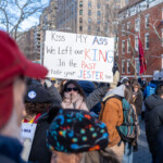 A protester at the 50 50 1 march in New York City on February 17, 2025, holds up a handmade sign with a defiant message that reads: "KISS MY ASS. We left our KING in the PAST, take your JESTER too." The bold lettering alternates in colors, emphasizing "ASS" in blue, "KING" in red, "PAST" in red, and "JESTER" in blue. The woman holding the sign is bundled up in a gray winter coat, beige scarf, and sunglasses, standing amidst a crowd of demonstrators. The protest took place in Washington Square Park, with a backdrop of historic red-brick buildings and New York’s urban landscape. Attendees are seen wearing warm clothing, hats, and scarves on a chilly winter day, engaging in political activism.