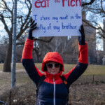 A protester at the 50 50 1 march in New York City on February 17, 2025, holds up a provocative sign that reads: "eat the rich. I store my meat in the belly of my brother." The lettering is bold, with "eat the rich" in red and the rest of the text in blue. The individual is dressed for the cold weather, wearing a red and navy blue insulated jacket with a hood up, black gloves, and white-framed sunglasses. A backpack is strapped to their shoulders. The setting is an outdoor park area, likely Washington Square Park, with leafless trees and an iron fence visible in the background. Other demonstrators are scattered in the vicinity, participating in the protest against economic inequality and political corruption.