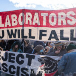 At the 50 50 1 march in New York City on February 17, 2025, a large group of protesters gathers in Washington Square Park holding striking banners and signs condemning fascism and political collaboration. The most prominent banner, in red and white, reads "COLLABORATORS YOU WILL FALL," while another below it states "REJECT FASCISM" alongside a snarling black wolf illustration. The crowd, dressed in winter coats, hats, and masks, stands united, holding signs with additional messages, including a handwritten one that says "Do unto others as you would have them do unto you." The atmosphere is resolute as demonstrators express their stance against authoritarianism and political alliances perceived as harmful. The backdrop features the park’s landmarks, city buildings, and a bright winter sky, with sunlight illuminating the banners.