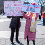 Two demonstrators at the 50 50 1 march in Washington Square Park, NYC, on February 17, 2025, hold bold protest signs calling for economic and political resistance. The person on the left wears a dark brown puffer coat and black boots, holding a large handmade sign that reads:
"BOYCOTT THE BILLIONAIRES"
It lists companies such as Trump Tower, Amazon, WWE, Tesla, SpaceX, Boeing, Meta, Uber, Exxon, Johnson & Johnson, and Uline, with the phrase "HIT 'EM WHERE IT HURTS!!!" written in red.
The person on the right is dressed in a long pink puffer coat, black boots, and a pink beanie, carrying a yellow polka-dotted tote bag. Their sign states:
"AMERICA FOUGHT A KING BEFORE, REPUBLICANS HAVE CROWNED ONE"
with additional messages: "NO KINGS, NO DICTATORS, NO TRUMP."
The protest takes place under a bright winter sky, with the Washington Square Park arch and surrounding buildings visible in the background. Other protesters carrying signs and banners fill the park, reinforcing the atmosphere of resistance.