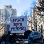A group of demonstrators at the 50 50 1 march in New York City on February 17, 2025, gather in the streets, holding up various protest signs. One prominently displayed sign reads "IMPEACH BOTH PRESIDENTS" with a red downward arrow and jagged lettering, symbolizing dissatisfaction with current leadership. The protesters, dressed in winter clothing, including knit beanies and gloves, stand together in solidarity against political figures. The background features tall residential buildings and bare trees, indicating the winter season. Other signs, including a U-Haul box repurposed into a protest sign, can be seen, adding to the grassroots energy of the demonstration.