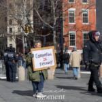An older protester stands in Washington Square Park in New York City on February 17, 2025, holding a handmade cardboard sign that reads, "CORRUPTION IS TYRANNY," with the word 'IS' highlighted in red. Dressed in a green coat, gloves, and sneakers, they maintain a quiet but determined stance in a public space known for activism.
In the background, two NYPD officers stand near metal barricades, observing the protest activity. Other demonstrators and pedestrians, bundled in winter clothing, walk past the scene, engaging in conversations. The setting is framed by the Washington Square Arch and the historic red-brick buildings of Greenwich Village, adding depth and a sense of place to the moment.
This image captures a powerful statement on political accountability and resistance at the 50 50 1 march, where citizens gathered to voice concerns over governance, democracy, and corruption. The solitary figure with the bold message highlights the power of individual activism amid a larger collective movement.
