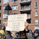 A demonstrator at the 50 50 1 march in New York City on February 17, 2025, holds up a handmade sign with a clear statement of resistance. The sign reads:
"I STAND IN OPPOSITION TO THIS ADMINISTRATION. IF YOU, TOO, FEEL THIS OPPOSITION WITHIN YOU, STAND WITH ME. STAND WITH ME. STAND WITH ME."
The individual, dressed in a gray winter coat, grips the sign firmly, elevating it above their head. Their face is partially obscured, but their stance conveys determination and solidarity. The protest takes place in an urban setting, with a red brick apartment building and balconies in the background, showing the presence of residents observing the demonstration from their homes. The crowd around the protester consists of individuals bundled up against the cold, including people wearing colorful hats and winter coats.
The moment captured highlights the power of peaceful protest and individual voices uniting against political leadership. The phrase "STAND WITH ME" repeated on the sign emphasizes a call for collective action.