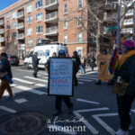 A protester stands in the middle of a crosswalk during the 50 50 1 march in New York City on February 17, 2025, holding a sign with a historical reference to democracy. The sign reads:
"We are a nation OF the PEOPLE, BY the PEOPLE, FOR the PEOPLE. Remember what the PEOPLE did to King George."
The message invokes the American Revolution, drawing a parallel between past struggles for democracy and the present. The protester, dressed in a dark winter jacket, gloves, and a hat, appears determined as they hold the sign firmly.
To the right, another demonstrator in a pink knit hat and yellow scarf holds a rainbow flag with a heart design, adding a LGBTQ+ solidarity element to the scene. In the background, a Spectrum News 1 van is parked on the side of the street, documenting the event. Additional protesters, some holding signs and banners, are visible further down the block. One banner in the background reads "First Unitarian Church", indicating community and faith-based involvement in the demonstration.
The urban environment, with brick apartment buildings, a police surveillance camera, and street signs, reinforces the city setting. The composition captures the intersection of historical references, modern political activism, and diverse causes.