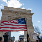 A large American flag waves high in the air in front of the Washington Square Arch in New York City during the 50 50 1 march on February 17, 2025. The bright blue sky and sunlight illuminate the historic marble arch, a symbol of freedom and resilience, as demonstrators gather for the protest.
In the foreground, a man in sunglasses and a beanie holds the flagpole, raising it with both hands. Other protesters are seen nearby, some holding signs, including one that reads "Veterans Say No to Fascism", reinforcing the march’s anti-authoritarian and pro-democracy message. Participants are bundled in winter clothing, indicating the event is taking place in cold weather.
The New York City skyline, including One World Trade Center, is visible in the background, further emphasizing the patriotic and civic themes of the demonstration. The flag, a powerful national symbol, serves as a focal point in this image, representing the ideals of justice, democracy, and resistance that many at the march are advocating for.