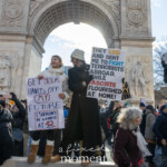 Two protesters stand on the base of the Washington Square Arch in New York City during the 50 50 1 march on February 17, 2025, holding bold signs addressing issues of youth concerns and military service contradictions. The woman on the left, dressed in a white coat and beige boots, holds a handmade sign that reads: "Get your hands off my future. I shouldn't be worried at 12." Her message speaks to youth advocacy, likely referencing political instability, climate anxiety, or social issues affecting younger generations.
Next to her, another protester in a long black coat, hat, and gloves holds a sign stating: "They lied and sent me to fight terrorists abroad, while fascists flourished at home!" This message strongly criticizes government policies and perceived domestic threats to democracy, a common theme at the demonstration.
Surrounding them, a large crowd of protesters in winter attire holds additional signs, with one partially visible sign in the crowd mentioning "Nazi" references. A participant on the right, wearing a red wig and face covering, holds up a smartphone, possibly recording the event. The Washington Square Arch looms in the background, emphasizing the historical and symbolic weight of the protest’s location.
This image captures the passion and urgency of the demonstrators advocating for justice, democracy, and civil rights.