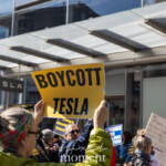In this photograph from the Picket Tesla protest held on March 1, 2025, at the Tesla Showroom in Manhattan (860 Washington Street, NYC), a demonstrator holds up a bold yellow sign reading "BOYCOTT TESLA" in large black letters. The person holding the sign is wearing a lime-green quilted jacket and has a colorful patterned headband securing their gray hair in a ponytail. Glasses rest on their face as they raise the sign high, their fingers gripping the edges firmly.
In the background, other protesters can be seen holding various signs with strong messages, including one partially visible reading "Those who remain silent will be as complicit as..." and another stating "Billionaires are killing you." The demonstration is taking place outside a Tesla showroom, clearly marked by the large glass facade and the "TESLA" logo on a sign near the entrance. The protesters are actively engaging, with some speaking and others raising their signs. A mix of individuals, bundled up in jackets and scarves, suggests the event is occurring during the colder months.
The event is likely part of a coordinated action against Tesla, possibly due to labor concerns, environmental issues, or political affiliations of its leadership. While no specific organization is named in the image, similar protests are often linked to labor unions, environmental activists, or political groups.