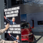 At the Picket Tesla protest organized by Rise and Resist (@risenresistnyc) on March 1, 2025, outside the Tesla Showroom in Manhattan (860 Washington Street, NYC), a demonstrator presses a bold black-and-white sign against the glass window of the store. The sign features a digitally altered image of Elon Musk in a Nazi-like salute, with the text:
"THIS BILLIONAIRE IS KILLING OUR GOVERNMENT"
"THIS BILLIONAIRE STOLE YOUR SOCIAL SECURITY NUMBER"
The protester, whose hands are visible gripping the edges of the sign, is wearing a beige jacket. The Tesla showroom interior is visible through the window, featuring a sleek red Tesla vehicle parked inside, with promotional signage that reads "Go Anywhere." The glass reflects the outside environment, including buildings and trees, blending the imagery of the protest with the luxury electric vehicle inside.
This demonstration, led by Rise and Resist (@risenresistnyc), is part of a broader movement against billionaire influence, corporate power, and the political and economic policies associated with figures like Elon Musk. The protesters make a striking visual statement by juxtaposing their anti-billionaire message directly against Tesla's branding inside the showroom.