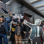 At the Picket Tesla protest organized by Rise and Resist (@risenresistnyc) on March 1, 2025, outside the Tesla Showroom at 860 Washington Street, NYC, activist Jay W. Walker and other demonstrators hold provocative signs condemning Elon Musk and billionaire influence.
In the foreground, Jay W. Walker, a well-known activist, raises a sign that repeatedly says "NAZI" alongside an edited image of Elon Musk in a salute-like pose. He wears a blue quilted jacket, dark jeans, a black t-shirt, and a cap adorned with buttons and pins. His mouth is open, as if leading chants or shouting slogans.
To his right, a protester in a dark blue jacket and scarf holds another sign that reads:
"THIS IS THE IMMIGRANT WHO TOOK YOUR JOB."
This sign satirizes anti-immigrant rhetoric to criticize Musk’s corporate policies.
In the center of the image, a life-size cardboard cutout of Elon Musk dressed in a suit and raising his arm is prominently displayed, reinforcing the protest’s visual impact.
Other demonstrators in the background hold additional signs, including one that states "DOGE IS A CRIMINAL ENTERPRISE", while another partially visible sign criticizes "SHADOW MONARCHS". A protester on the right wears a Superman-inspired LGBTQ+ pride t-shirt and a black-and-white keffiyeh scarf, engaging in discussions with fellow attendees. Many participants wear warm clothing and sunglasses, suggesting a chilly day.
The Tesla showroom’s reflective glass doors serve as a backdrop, merging the corporate space with the high-energy demonstration. The protest, led by Rise and Resist (@risenresistnyc), calls out Elon Musk’s influence, corporate greed, and wealth inequality in a visually striking and confrontational display.