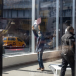 At the Picket Tesla protest organized by Rise and Resist (@risenresistnyc) on March 1, 2025, outside the Tesla Showroom at 860 Washington Street, NYC, a tense moment unfolds as a security guard in black clothing and beige boots gestures toward a protester inside the showroom. The protester, wearing a blue jacket and a face mask, holds a small American flag in one hand and a black sign with white lettering that reads, "THIS BILLIONAIRE IS DESTROYING OUR COUNTRY" in the other.
This striking image is captured from the outside looking in, with reflections of protesters and their signs merging with the showroom's sleek interior. The Tesla humanoid robot display is visible in the background, symbolizing the company's futuristic branding, while the presence of protesters inside contrasts sharply with the polished corporate setting.
Outside, reflections of demonstrators holding yellow protest signs with messages against Elon Musk, corporate greed, and economic injustice can be seen on the showroom's glass windows, blurring the lines between activism and commerce. The New York City streetscape, complete with a yellow taxi and brick buildings, is also visible in the reflection, grounding the protest in its urban setting.
The Rise and Resist (@risenresistnyc) demonstration highlights growing concerns about billionaire power, political influence, and the role of corporations like Tesla in shaping the economy and government policy. The presence of security inside the showroom underscores the tension between activists and corporate interests.