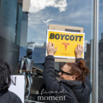During the Picket Tesla protest organized by Rise and Resist (@risenresistnyc) on March 1, 2025, outside the Tesla Showroom at 860 Washington Street, NYC, a woman in a black puffer jacket and sunglasses affixes a bold “BOYCOTT TESLA” sign onto the showroom’s glass facade. The yellow and black sign prominently features the Tesla logo, symbolizing opposition to Elon Musk’s leadership and corporate practices.
In the reflection of the showroom glass, other demonstrators are visible, along with the robotic humanoid figure displayed inside the Tesla store, adding a stark contrast between technology and protest. Another protester, also wearing a black jacket, stands beside her holding a different sign, engaging in collective activism against corporate power.
The protest, organized by Rise and Resist (@risenresistnyc), criticizes billionaire control, financial corruption, and Musk’s political influence, calling for economic resistance through boycotting Tesla and its associated enterprises.